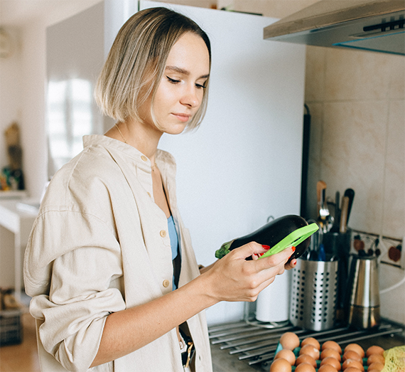 Woman with blond hair wearing a beige jacket using her phone in a kitchen.