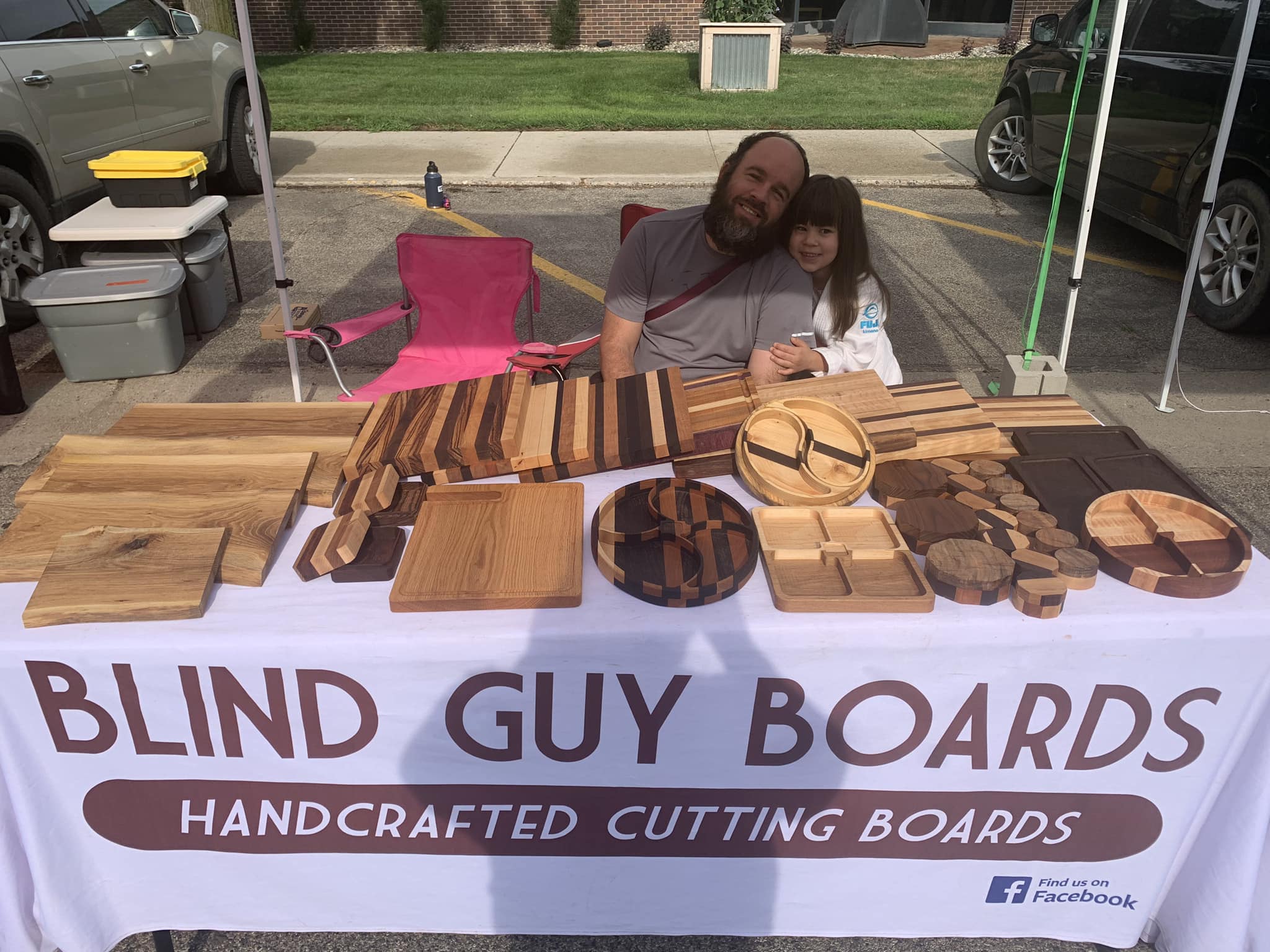 Ismael Collazo and his daughter sit behind a display table at an outdoor market, showcasing handcrafted wooden cutting boards of various shapes and designs. A banner on the table reads 'BLIND GUY BOARDS' with the tagline 'HANDCRAFTED CUTTING BOARDS,' along with a Facebook logo for more information.