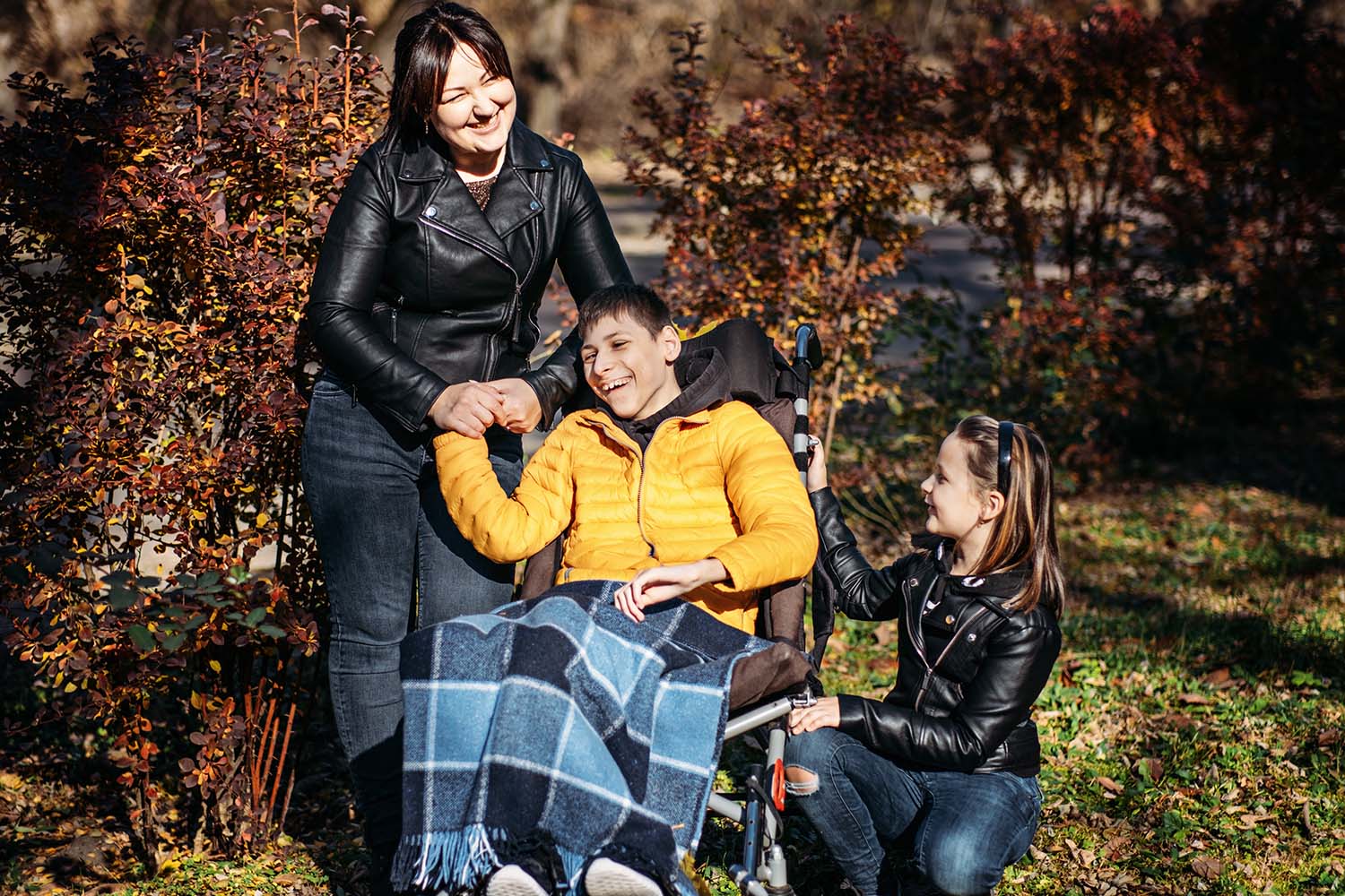 A mother stands next to her son in a wheelchair outside as her younger daughter kneels beside him. 