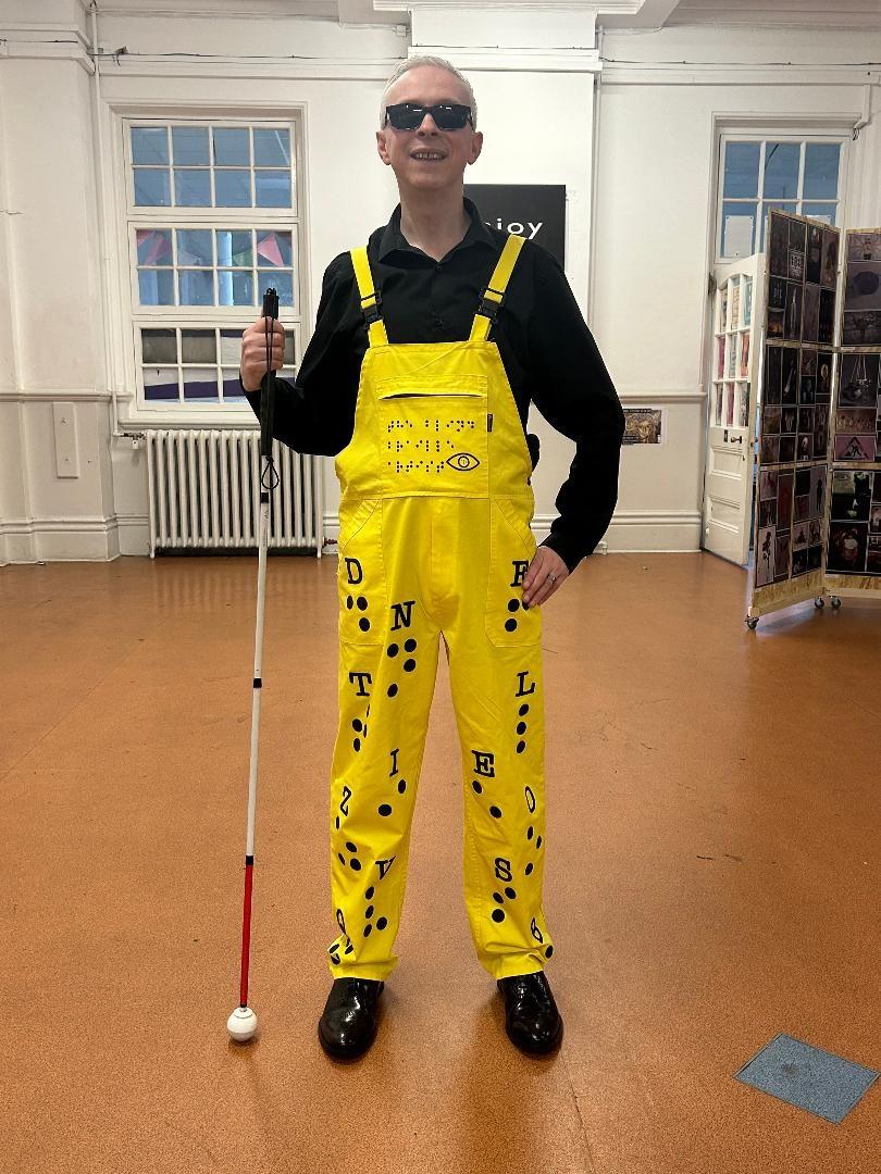 Clarke Reynolds stands in room holding his white can. He is smiling and wearing sunglasses and yellow overalls with braille on the pant legs.