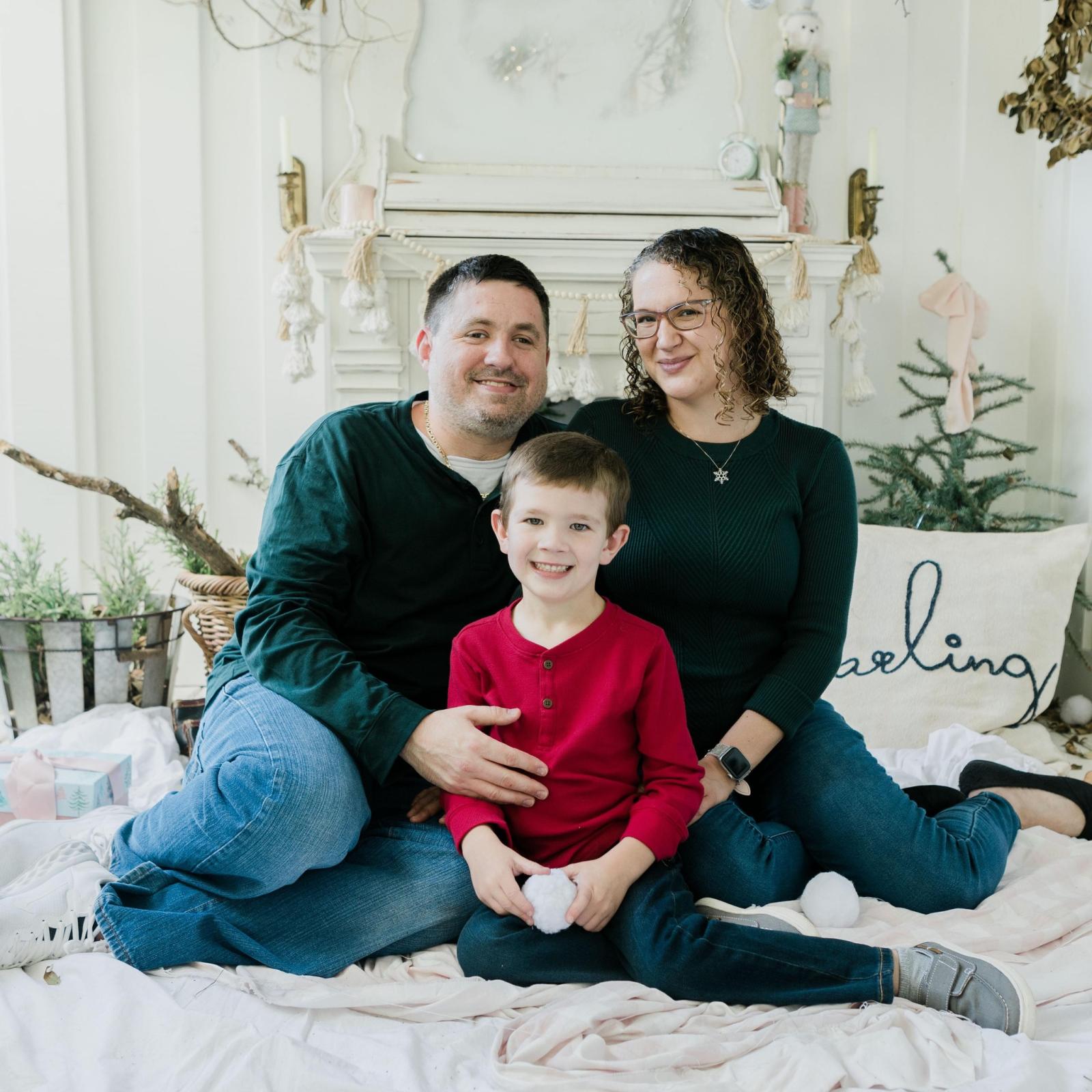 A family sitting together on a cozy white blanket in front of a decorated fireplace. The adults are wearing dark green tops and jeans, while the child in the center is dressed in a bright red shirt and dark pants. The background includes holiday-themed decorations such as greenery, a small tree, a basket, and a pillow with cursive writing. Soft lighting creates a warm, festive atmosphere.